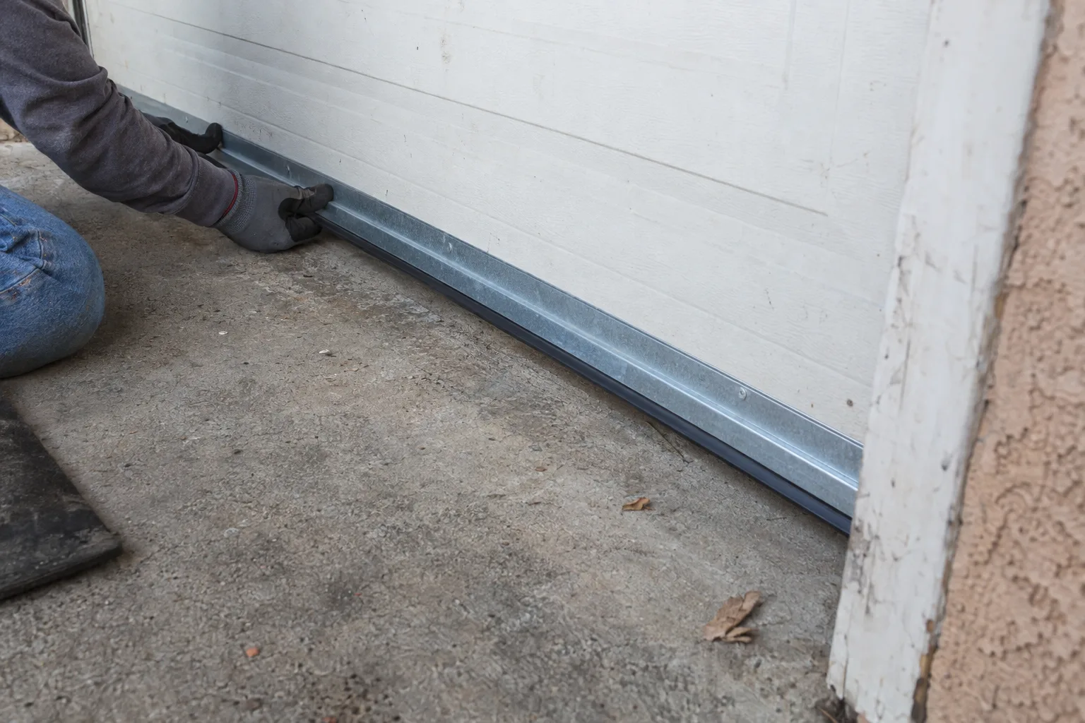 A gloved technician kneeling at the base of a residential roll-up garage door, pressing a new metal door bottom seal flush against a sun-bleached concrete driveway in an Los Angeles neighborhood