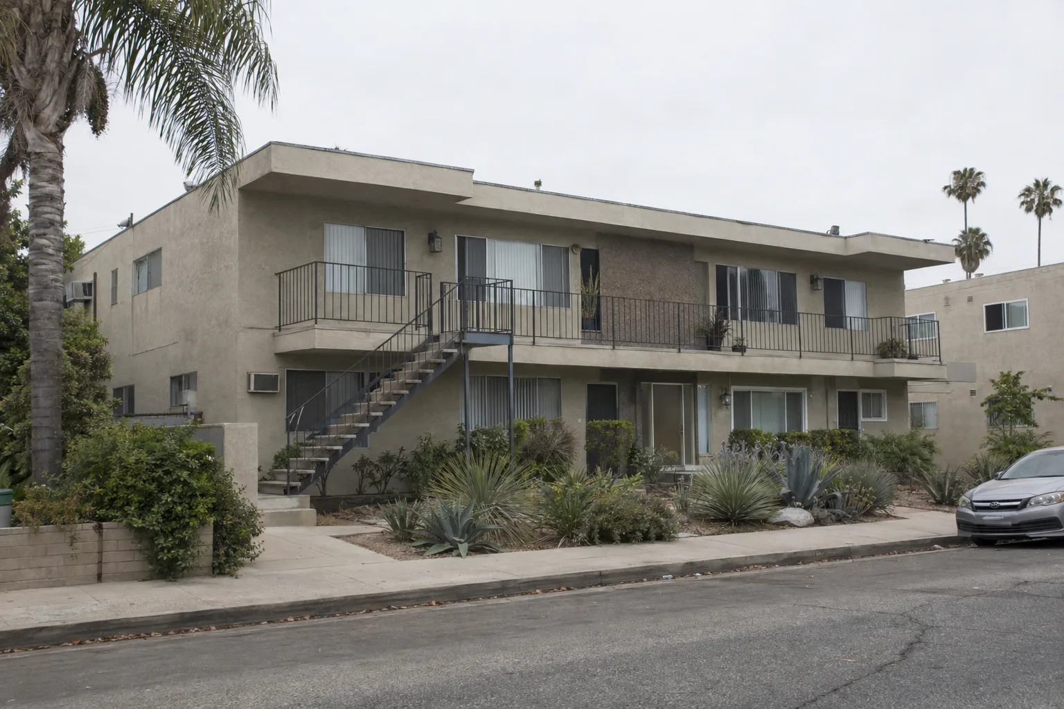 Two-story stucco apartment building from the 1970s on a residential street in the San Fernando Valley, with an exterior staircase, metal railings, and a palm tree nearby