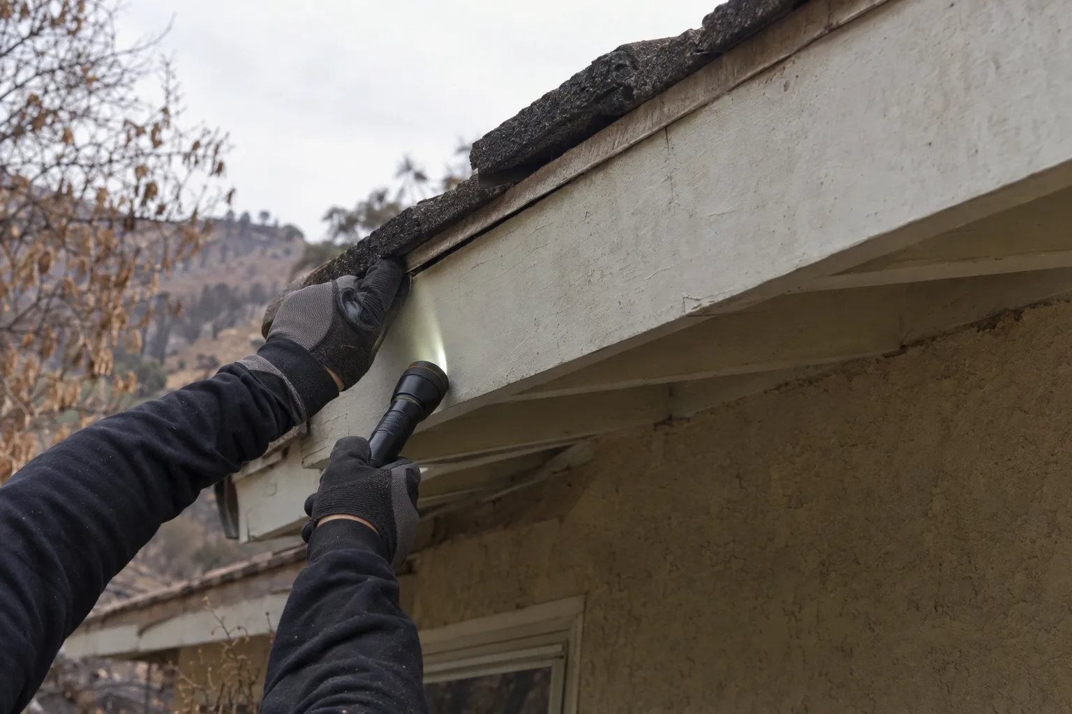 Gloved hands holding a flashlight examining a soffit gap along the roofline of a stucco Los Angeles home, with dry fire-scorched hillside vegetation visible in the background
