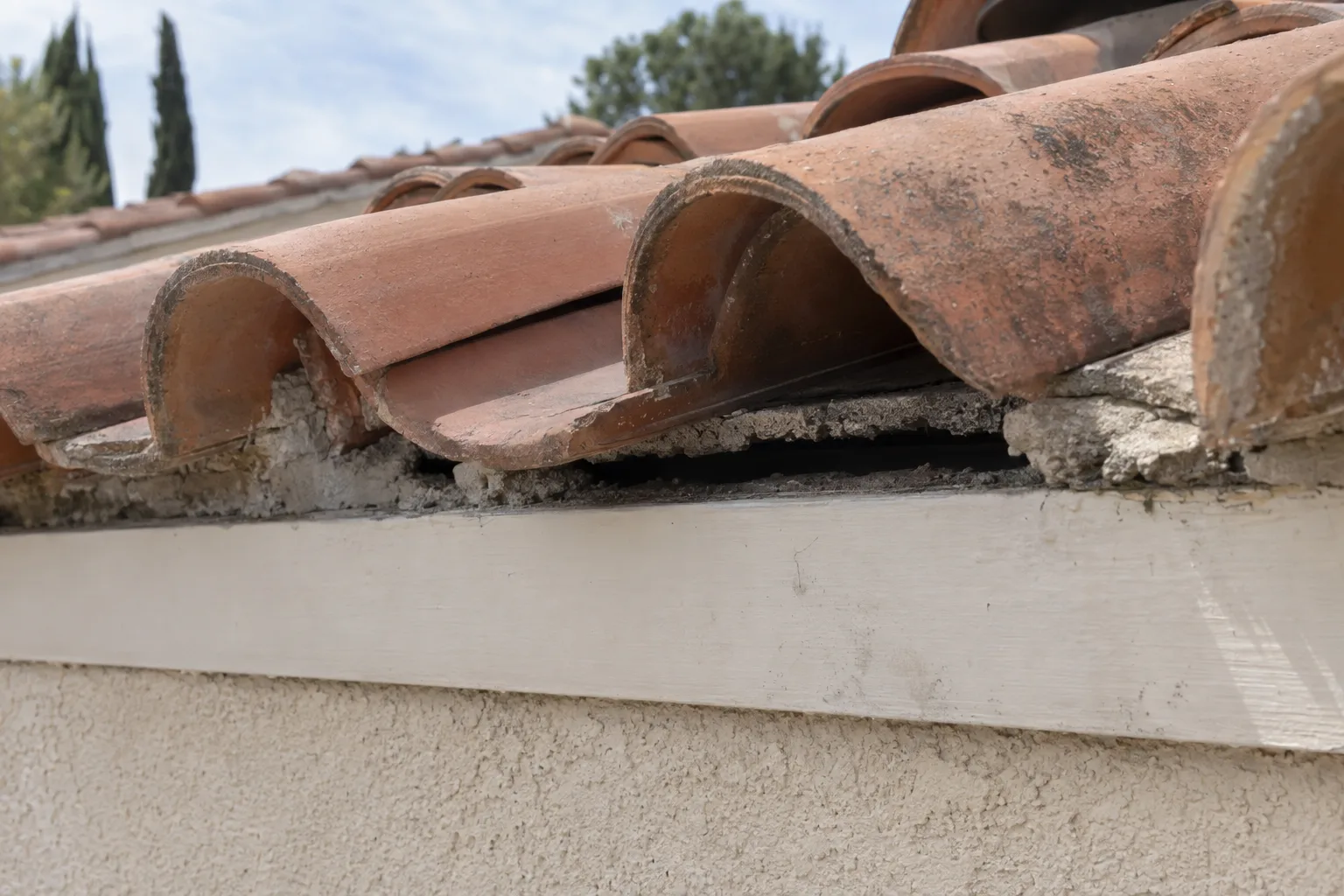 Close-up of a Spanish clay tile roofline on a stucco Los Angeles home showing an eroded bird-block gap at the eave, a dark void visible between displaced pan tiles and the fascia board