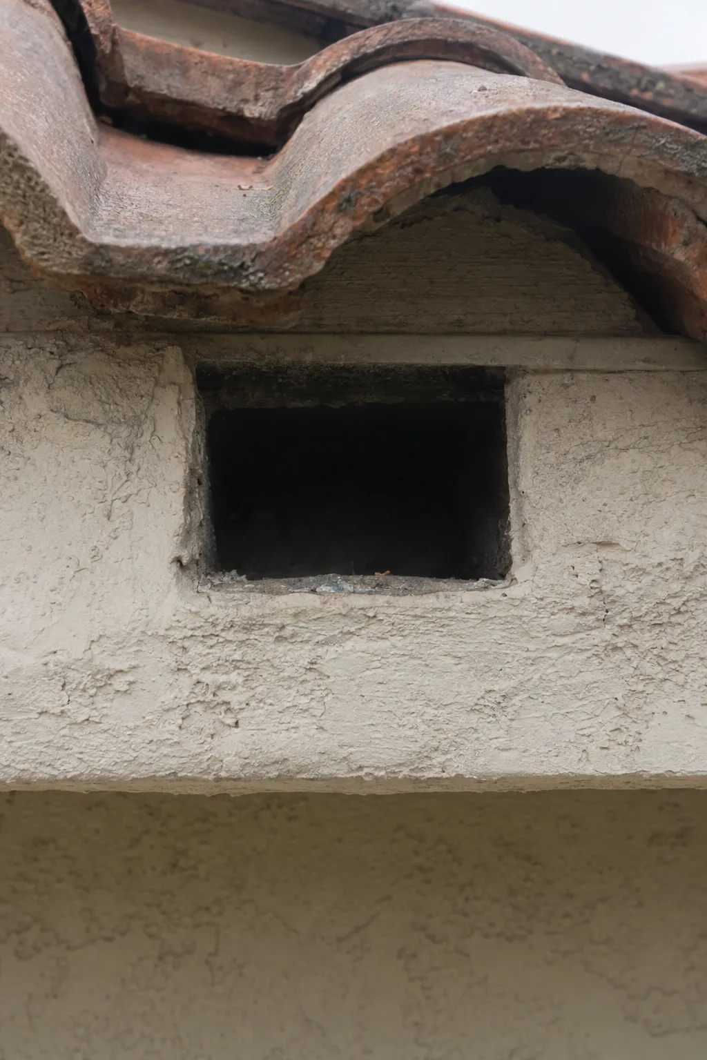 Open bird block gap beneath Spanish clay roof tiles on a stucco home, showing the dark unobstructed rectangular cavity at eave level