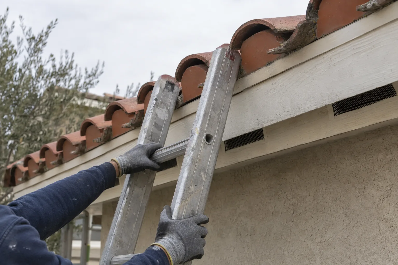 Gloved hands on ladder rungs at the eave of a Spanish clay tile roof, showing the roofline and open bird block openings on a stucco home in bright Los Angeles sunlight