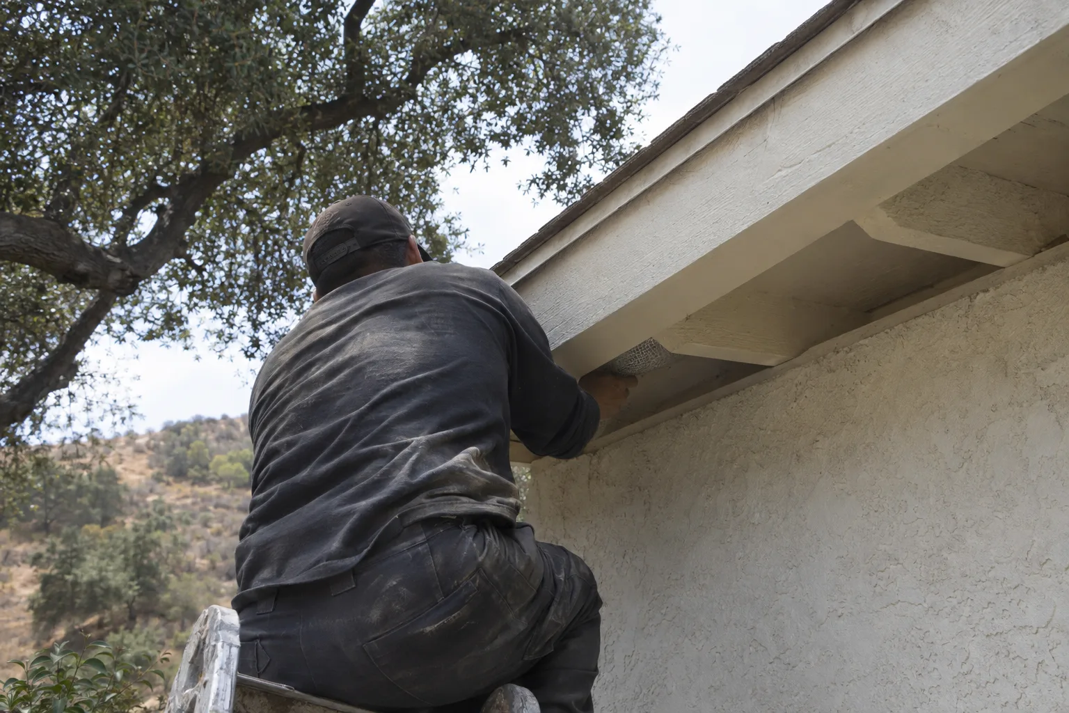 A worker crouching at the roofline of a white stucco Los Angeles home, pressing mesh material into a gap between the fascia and soffit, with dry California vegetation visible in the background